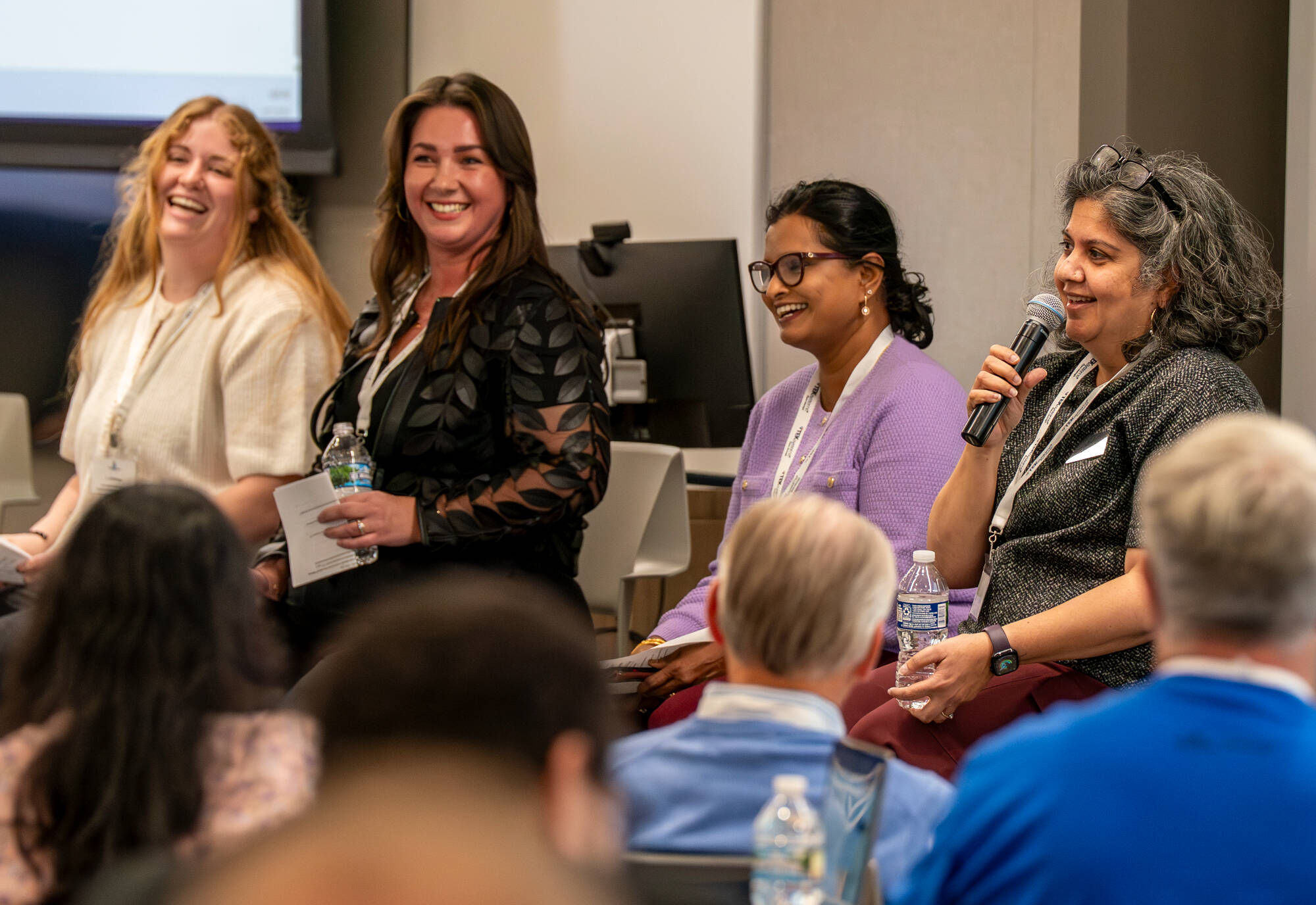 four women sit on tall chairs, person on right with microphone during a panel discussion; audience seated in front